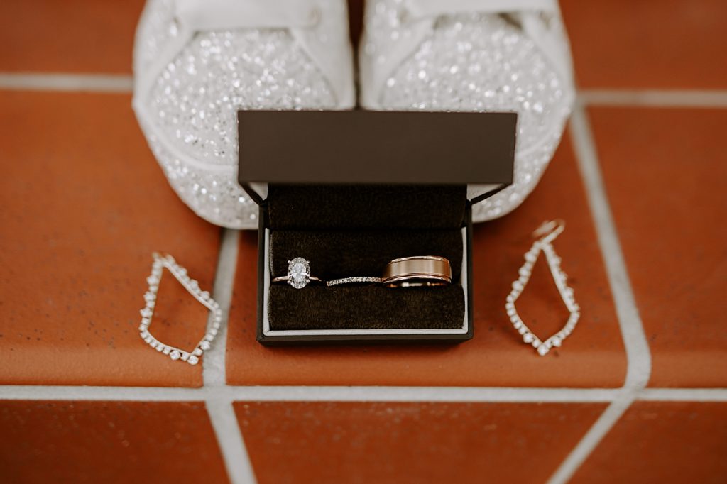 Detail shot engagement ring and wedding bands in box next to earrings by wedding shoes