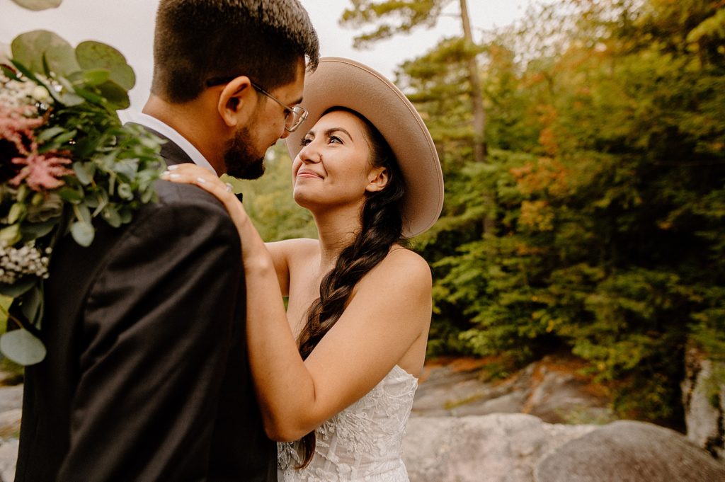 Bride with hat on with arms around Groom by outdoor forest