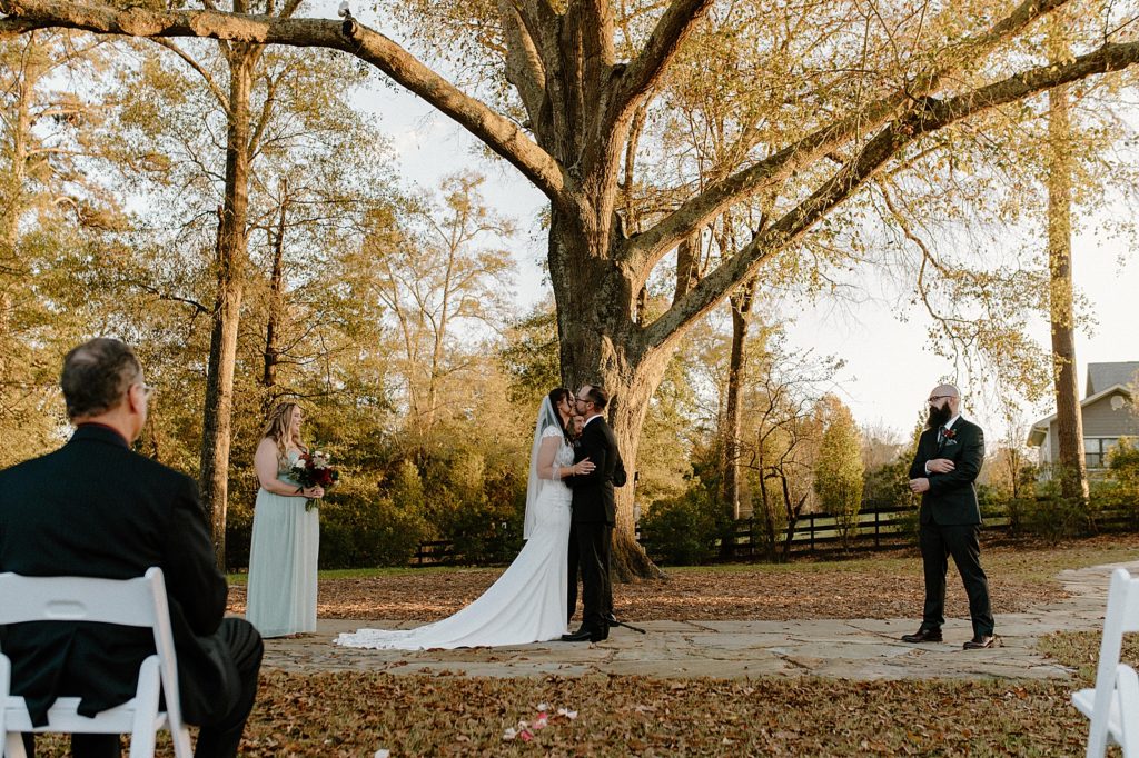 Just married Bride and Groom kissing at Ceremony by big autumn tree