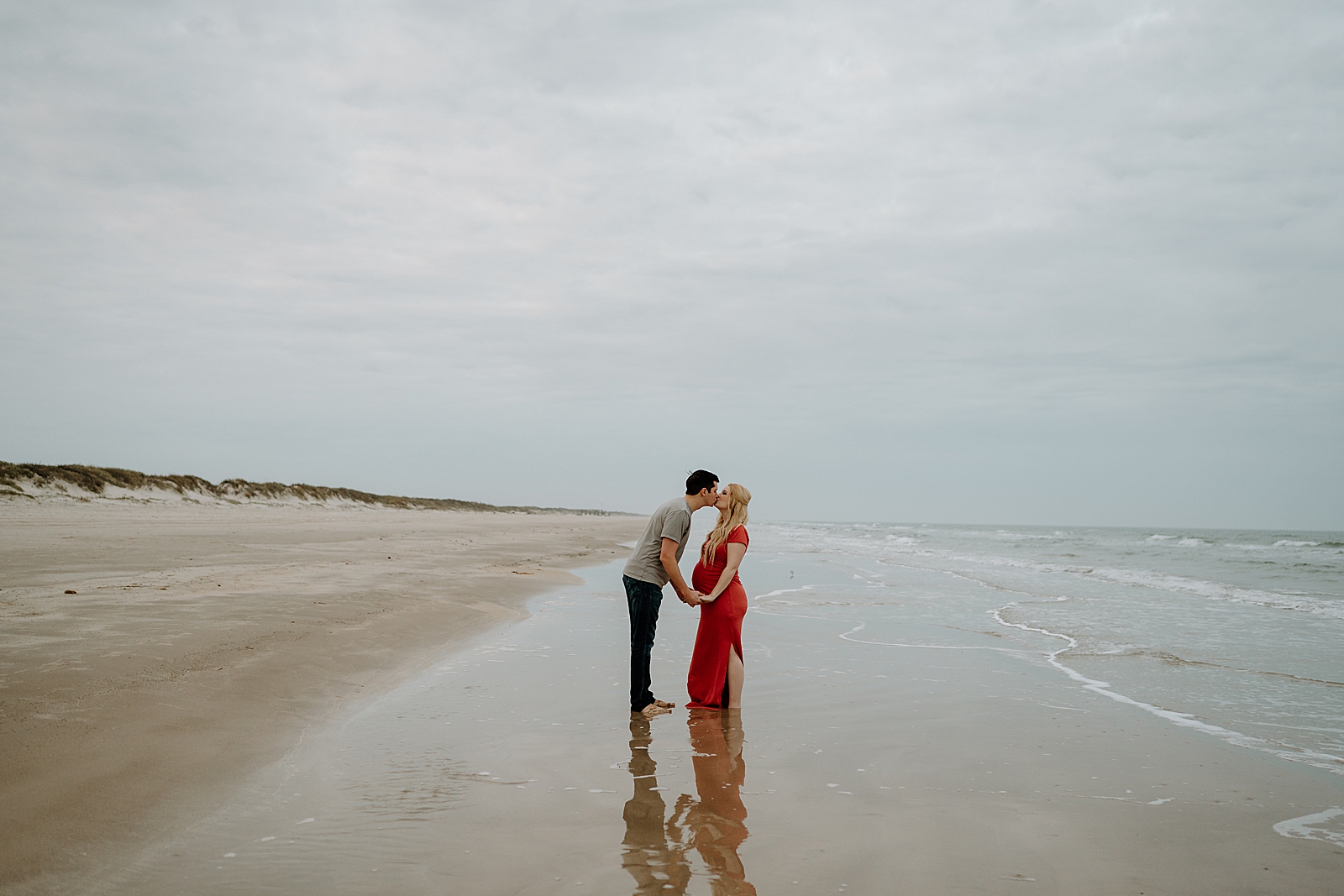 pregnant woman in red dress kissing spouse on beach