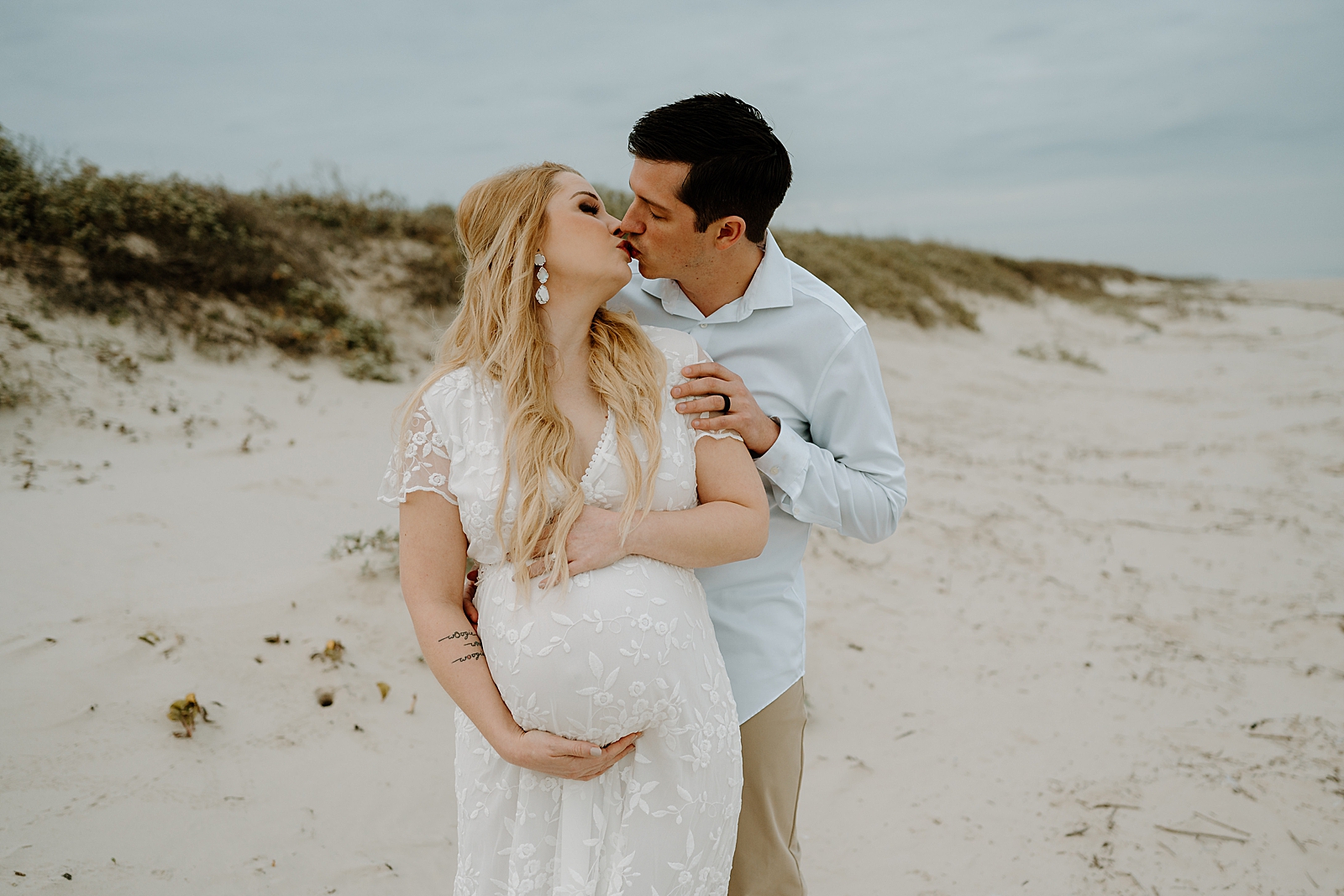 man in blue shirt kissing blonde woman in white dress holding belly bump on beach